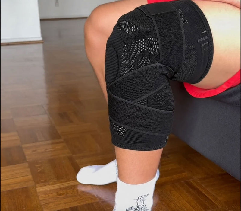 Person seated indoors wearing a black TEVANLY wraparound knee brace with adjustable straps.