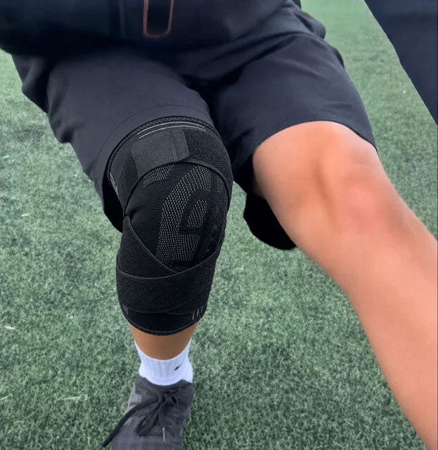 Close-up of a person wearing a black TEVANLY adjustable knee brace on artificial turf.