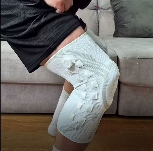 Person seated on a sofa wearing a white TEVANLY adjustable knee brace with side supports.